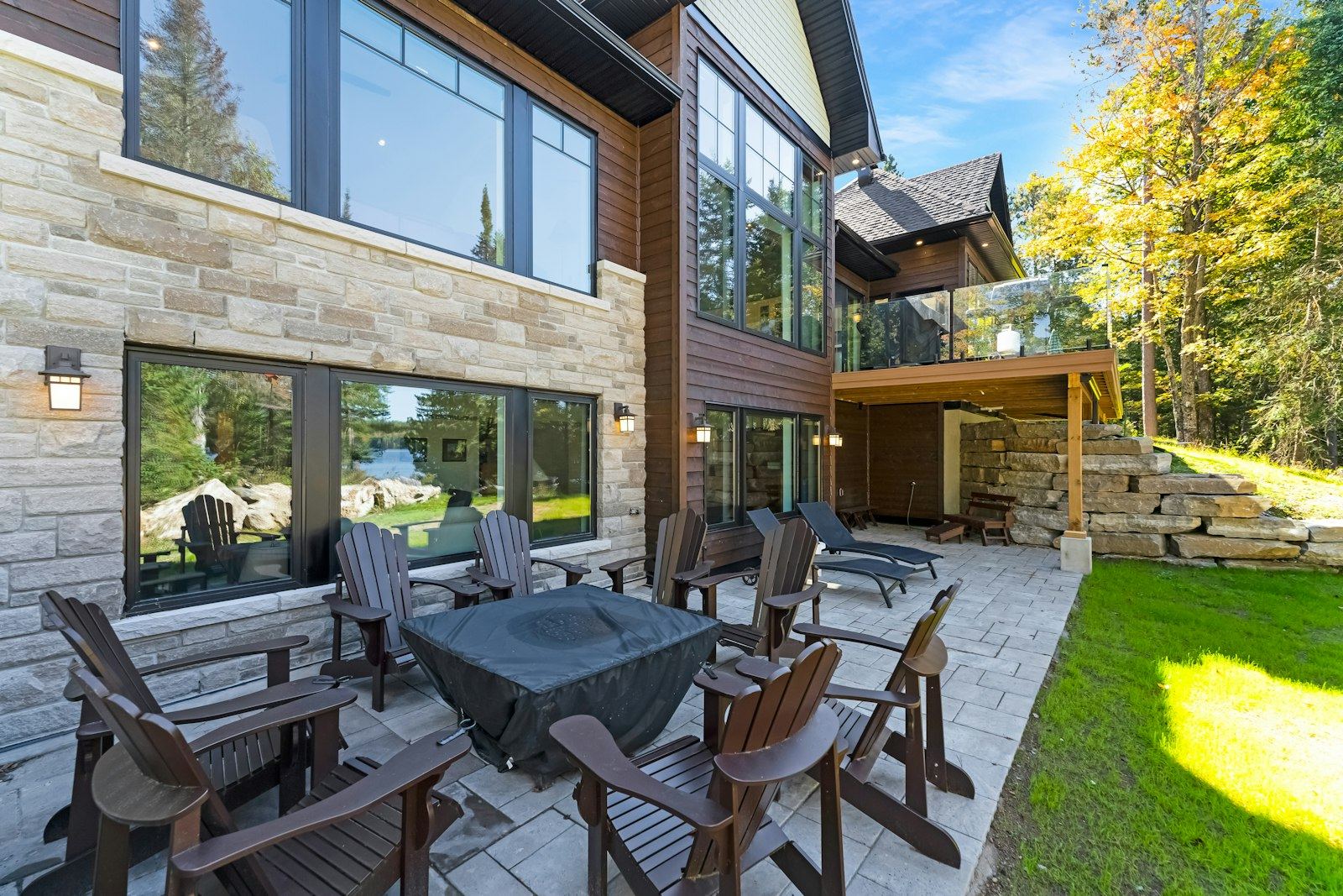 A stone paver patio with dark Adirondack chairs around a covered fire table beside a house